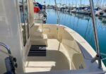Back deck of a white motor yacht at a marina, with seating and steps, calm blue water and moored boats in the background.