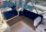 Lounge seating on a boat: tan cushions with dark blue back cushions, white captain chairs nearby, marina and water visible through clear panels.