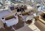 Cockpit of a motor yacht with a central steering wheel, dashboard gauges, and four white captain chairs; marina in the background.