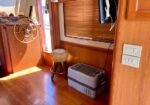 Wood-paneled boat cabin with a steering wheel on the left, a small woven stool, and a cooler on the floor by a window with blinds.