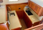 Compact boat galley with wooden cabinets, beige countertops, a small sink on the left, and a two-burner stove on the right, plus a ladder-step opening in the center.