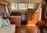 Interior of a yacht cabin showing a wood-paneled helm with steering wheel, kitchenette, and a beige sofa with blue pillows