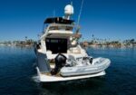 White luxury motor yacht with a gray inflatable dinghy mounted on the stern, in calm blue harbor water with a marina and palm trees in the distance
