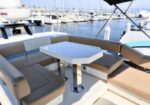 Yacht cockpit with beige cushioned seating around a white hexagonal table; marina with sailboats in the background.