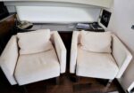Two cream-colored armchairs with matching cushions sit in a small living area on a dark wood floor, with a window shade behind and a shelf holding a hat and magazines nearby.