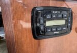 Black marine stereo head unit mounted on a wooden panel with display screen and labeled controls (Infinity). It includes volume, tune, mode, and presets.