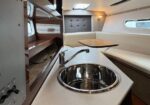 Interior of a boat galley with a stainless steel sink, curved faucet, and beige countertop; wooden shelves and a U-shaped seating area around a small table.