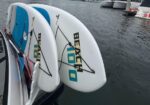 Two white boards (likely windsurf/surfboards) tied to a boat at a marina, with blue traction pads and cords, floating on calm water behind them.