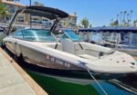 White and dark blue motor boat docked at a marina with the canopy raised, registration CF 2364 VW, moored beside other boats and a seawall with buildings and palm trees in the background.
