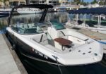 White and black speedboat docked at a marina, featuring seating, a small wooden table, and a black canopy; calm water and other boats visible in the background.