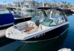 A dark blue motor boat docked in a marina with white interior seats and a small wooden table, calm water, and nearby houses.