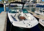 White motorboat docked at a marina with beige seating, a small wooden table, and a black canopy; calm water and waterfront homes in background.