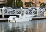 White center-console motorboat docked at a marina alongside a wooden pier, with a blue-gray water and parked cars in the background.