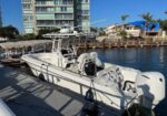 White motorboat docked at a marina along a wooden dock, with calm water and a blue sky, and a modern multi-story building and palm trees in the background.