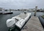 White motorboat docked at a wooden pier in a marina, with an outboard engine and seating area under an overcast sky.