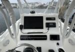 Helm of a white boat cockpit with a large navigation screen, radio, and control panels; marina buildings visible in the background.