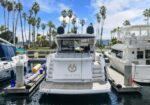 White luxury motor yacht docked at a marina, flanked by other boats with palm trees and blue sky in the background; flag on the stern and the name Veni Vici visible.