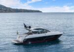 White luxury motor yacht cruising on calm blue sea with a rocky coastline and green hills in the distance.