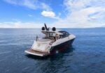 White luxury motor yacht cruising on calm blue water under a partly cloudy sky, viewed from the stern with outdoor seating area visible