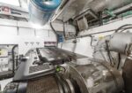 Engine room of a vessel with a large diesel engine, metal pipes, ducts, and control panels surrounding the machinery