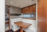 Interior of a boat cabin with white cushioned bench seating around a wooden table, porthole window, and warm wood cabinetry above. A doorway leads to a adjoining sleeping area.