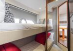 Cozy boat cabin with twin bunk beds, white linens and red pillows, a porthole window, and a doorway to another bunk area