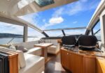 Sunlit yacht cockpit/lounge with white leather seating, wooden table, and helm area on the right, large panoramic windows and open sunroof showing blue sky and sea.
