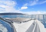 Deck of a white yacht with cushioned sun loungers on the bow, railing along the water, blue sea and sky ahead.