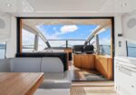 Interior of a luxury yacht with a seating area and wooden table, open doorway to the helm area with two captain chairs and a view of blue water and sky.