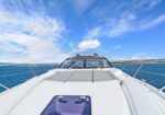View from the bow of a white yacht cruising on calm blue sea under a bright, partly cloudy sky. In the distance, a coastline is visible on the horizon.