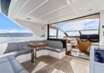 Luxurious yacht deck seating area with curved gray cushions, wooden table, and open doorway to the helm, ocean view in the distance.