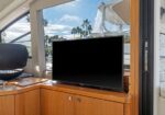 Flat-screen TV on a wooden cabinet inside a yacht, with large windows showing palm trees and blue sky outside.