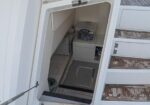Interior of a small boat cabin visible through an open hatch: a bunk bed to the left and a small storage cabinet with a clear plastic bin on top.