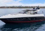 White and black luxury motor yacht cruising on calm blue water with a coastal town skyline in the background; sleek hull, tinted windows, and radar domes.