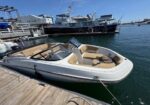 White motorboat with beige seating moored to a wooden dock, with larger yachts and a marina in the background.