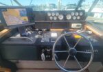 Captain's cockpit on a motorboat with a steering wheel, gauges, GPS/chartplotter, and throttle levers at the helm in a marina.