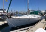 Sailboat moored at a marina on a sunny day, white hull tied to a wooden dock with other boats in the background.