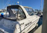 White motorboat named 'TWO BEST DAYS' moored at a marina with a beige canopy and blue sky above