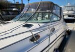 White motor yacht docked at a marina, bow close-up, with a tan canvas cockpit cover and clear windshield panels. View shows water and other boats nearby.