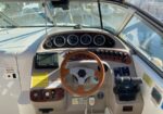 Boat helm cockpit with a wooden steering wheel, analog gauges, navigation screen, and throttle levers under a sunny cockpit canopy loadout.