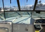 View from the boat's helm looking toward a marina, with the steering console, gauges, and navigation gear in the foreground and moored boats outside the windshield and blue sky above.