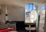 Interior of a small boat cabin with a cushioned bench, a small table, and an open doorway to the deck showing blue sky outside.