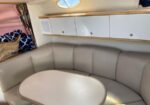 Boat cabin with a curved beige seating area around a rounded table and white overhead cabinets, plus a porthole window.