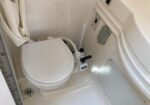 White marine toilet installed in a small boat head, with a manual pump handle and hoses visible in the surrounding compartment.