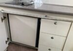 Under-counter kitchen scene with a white cabinet door open revealing a built-in appliance (likely a dishwasher) beneath a gray countertop, and white drawers with metal knobs to the right, plus a chrome rod along the front.
