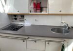 Small kitchen workspace with a two-burner stove on the left, sink on the right, gray countertop, and open cabinet with red cups inside.