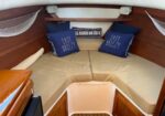 Boat cabin with a V-berth and beige cushions, two blue pillows reading 'Easy Breezy' and a back pillow saying 'All Hands on Deck', wood cabinets, and side nets.