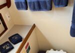 Blue towels with 'JOY' embroidered hang over a toilet in a small bathroom, with a toilet paper roll on the wall and folded JOY towels on a shelf nearby.