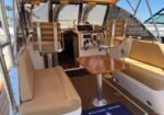 Sunlit boat cockpit with beige cushions, a wooden foldable table, steering wheel, and helm controls; blue 'Welcome Aboard' mat with an anchor.
