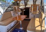 Boat cockpit with beige cushioned seating around a wooden pedestal table; two wooden captain chairs and an open helm in the background, under a canopy, with a 'Welcome Aboard' mat nearby.